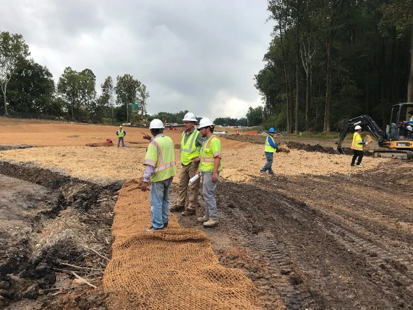 An image of three construction workers standing at a site where ground is being dug up and moved to prepare for a roadway expansion.
